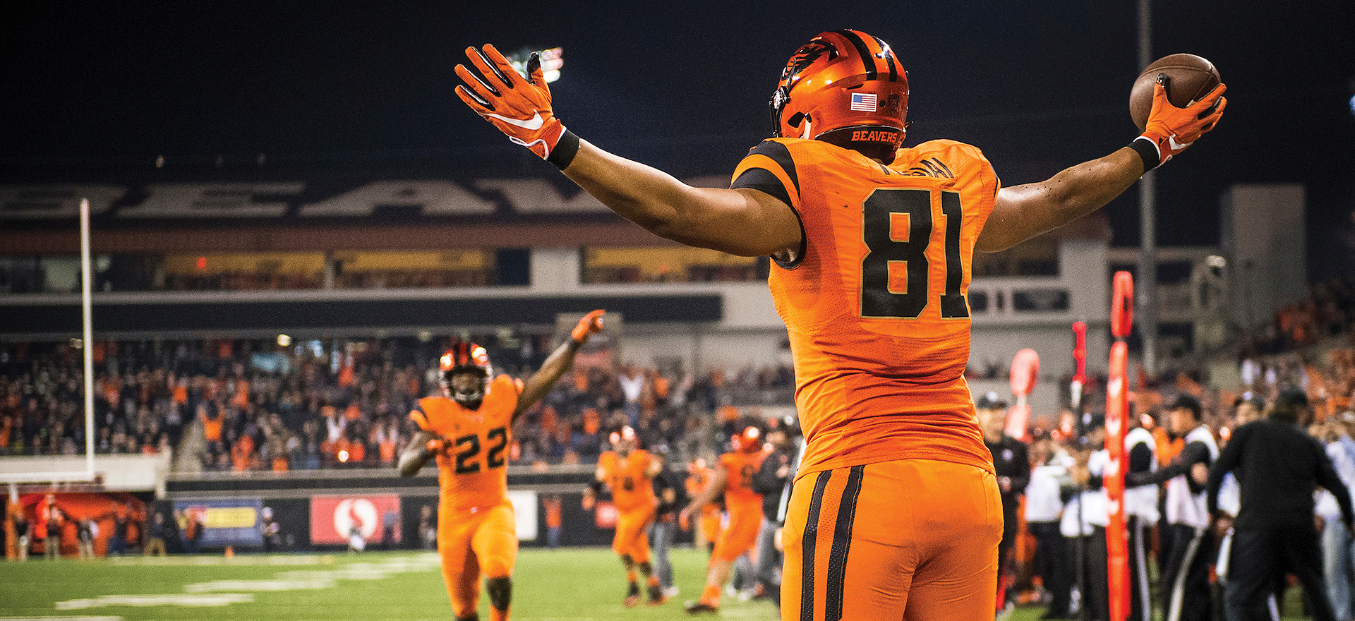 Football player on a football field during a game in orange jersey with back facing camera. His arms are raised up and it's night time.