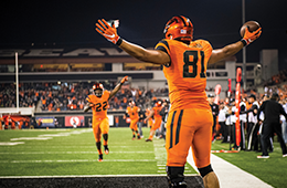 260x170_EventThumbnail_OSUAA-Football Football player on a football field during a game in orange jersey with back facing camera. His arms are raised up and it's night time.