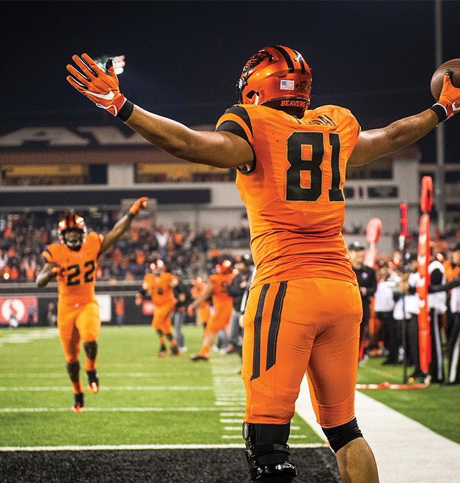 Football player on a football field during a game in orange jersey with back facing camera. His arms are raised up and it's night time.