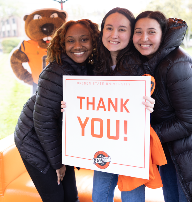 Three students holding a 'Thank You' sign and smiling for the camera on the MU Quad.