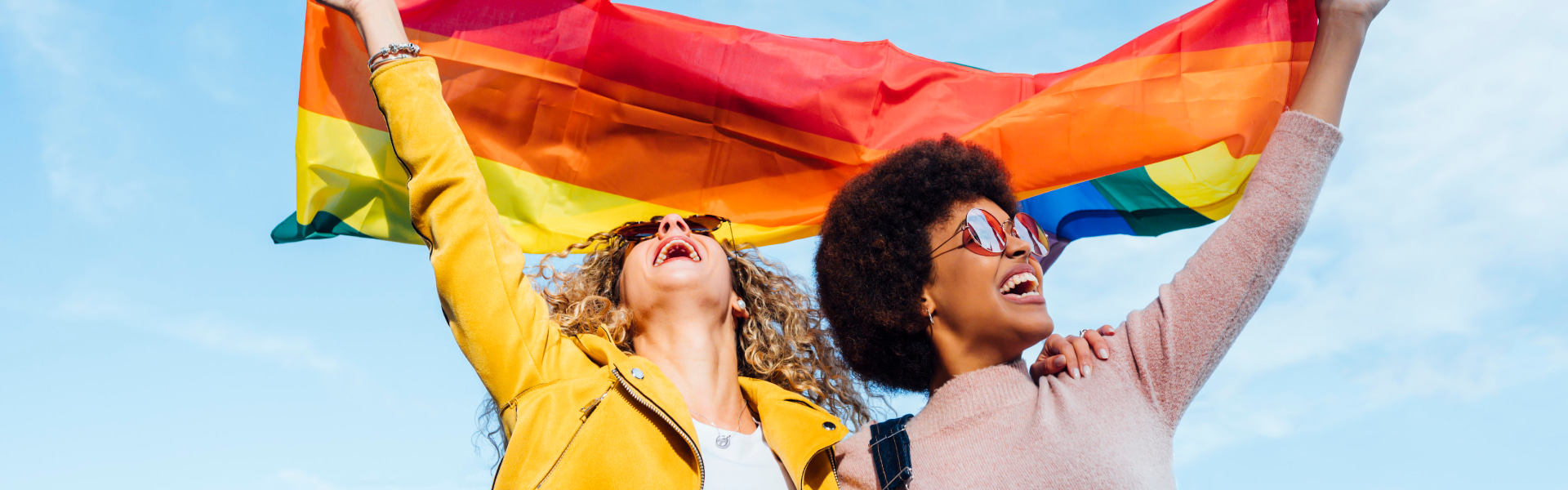 two women holding up a pride flag outdoors with a beautiful blue sky in the background