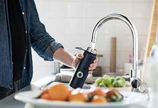 A person adding water from the sink to their black water bottle, with fruits including orange and limes on plates that are out of focus