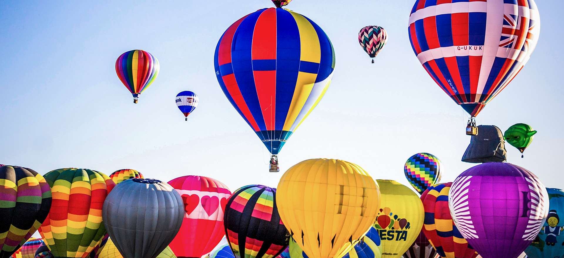A group of about 30 vibrant hot air balloons crowd together on the ground and in the air against a clear, blue sky.