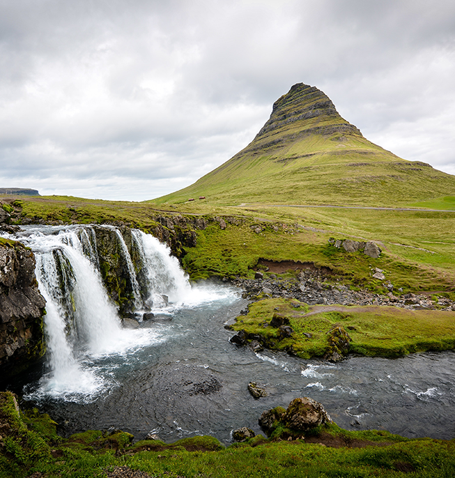 Overcast sky with small waterfall, river, and pointed green mountain.