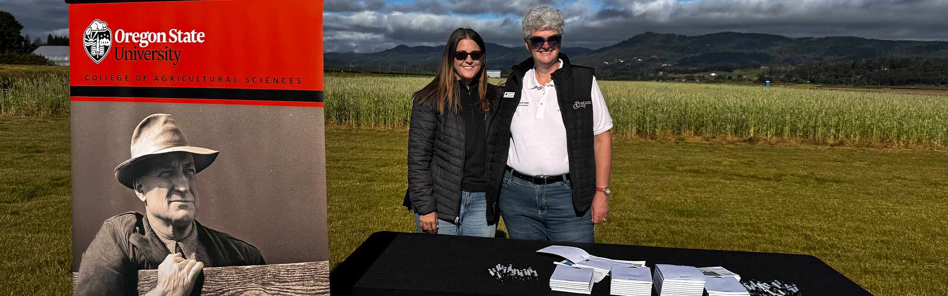 Two people stand behind an Oregon State University College of Agricultural Sciences table at an outdoor event, with an E.R. Jackman Friends and Alumni banner and farmland in the background.