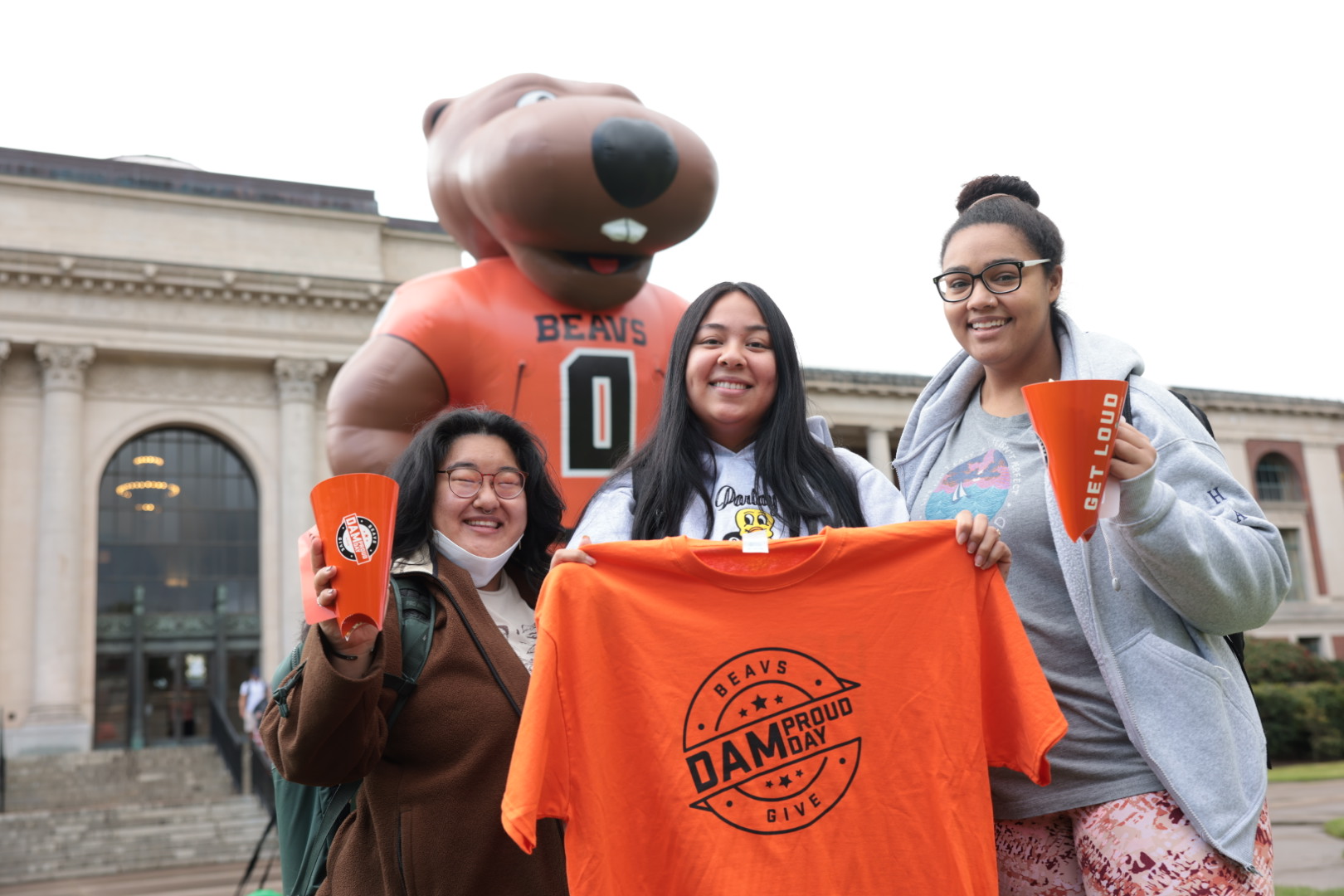 Students holding OSU swag including a Tshirt and cups outside of OSU campus with a blow up Benny behind