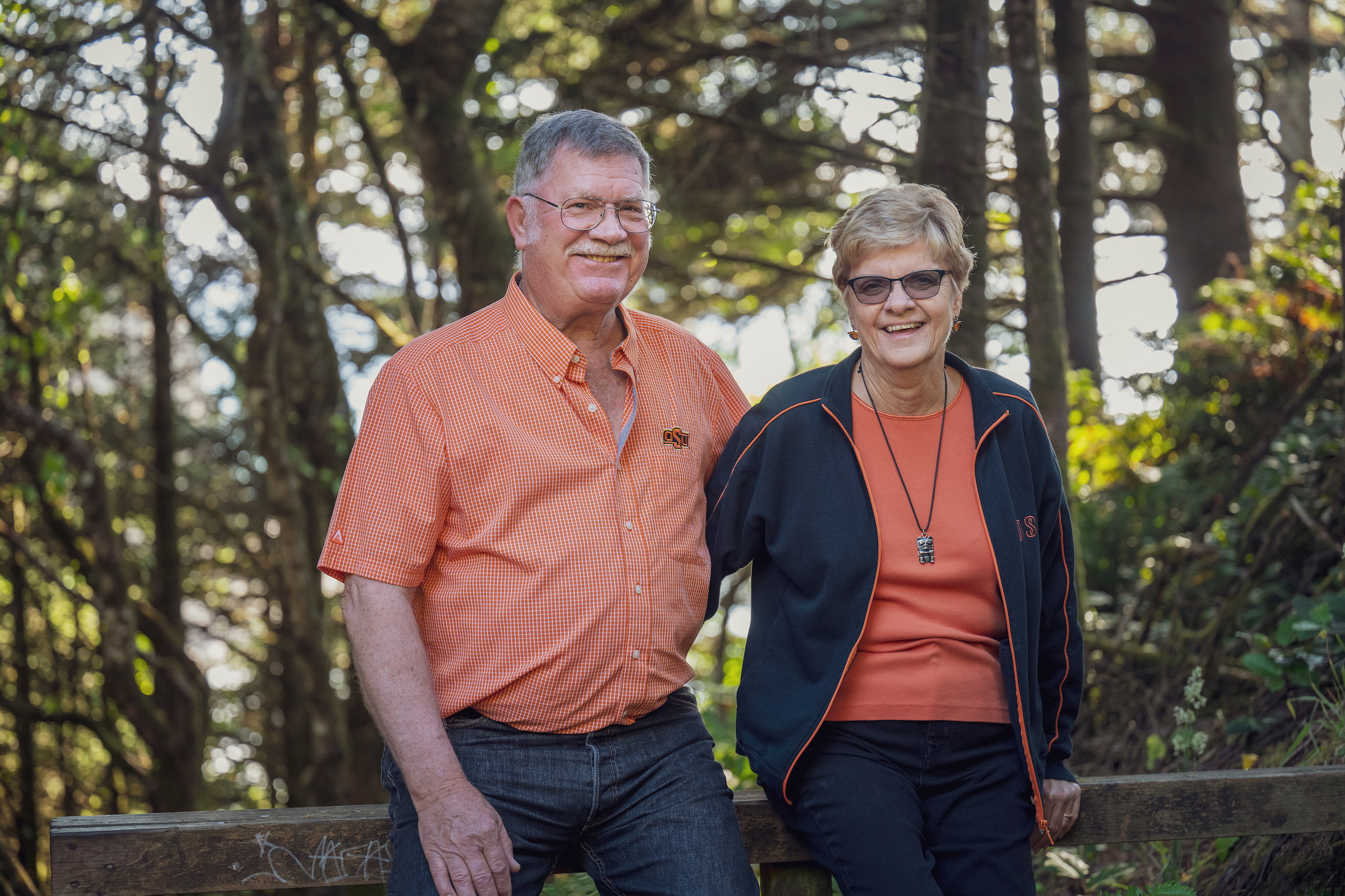 Nancy and Kent Searles wearing orange, background is of Oregon coast trees and you can see hints of the ocean behind the trees