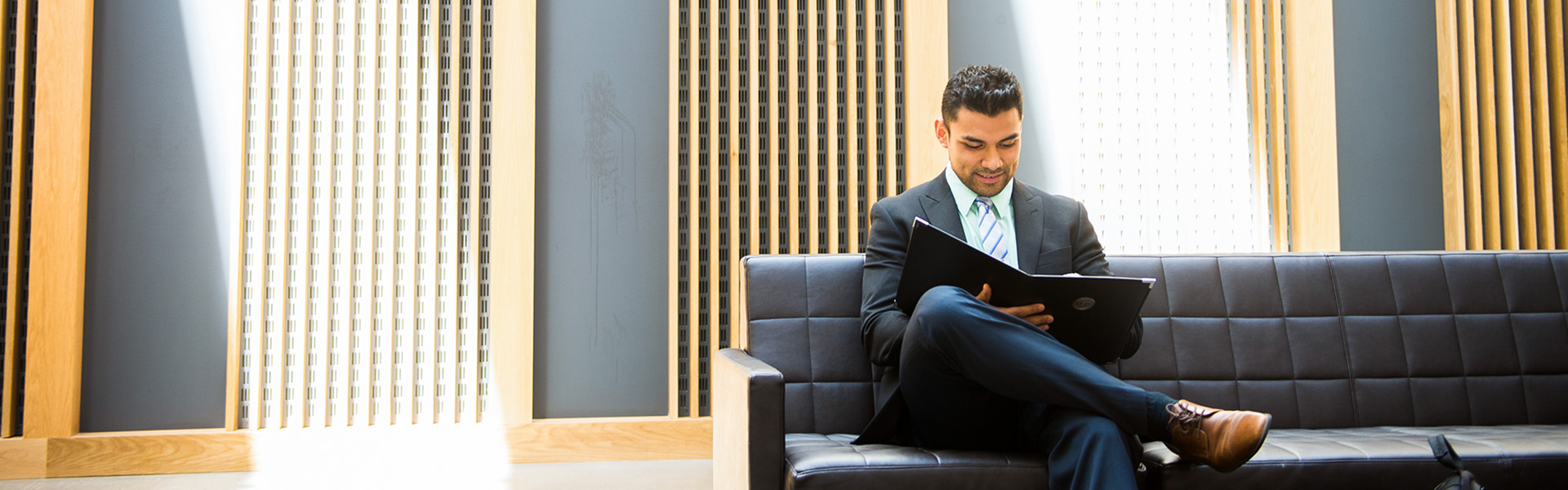 A student in a suit sits on a sofa looking over a binder with rays of sunlight beaming behind him.