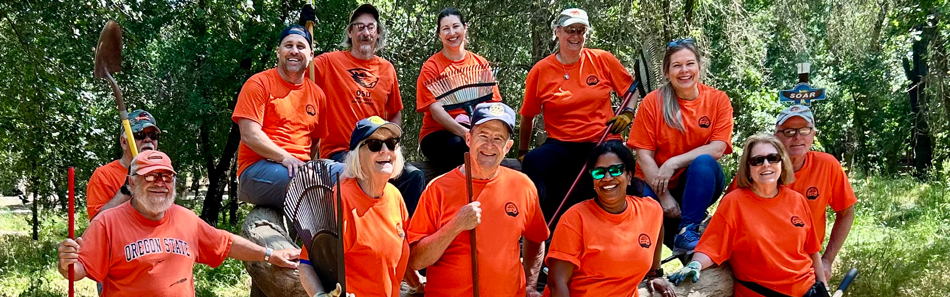 OSU volunteers in orange shirts hold wheelbarrows, rakes and other garden tools.