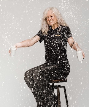 Person seated on a stool holding two beverage cans as liquid sprays around them in a studio setting.